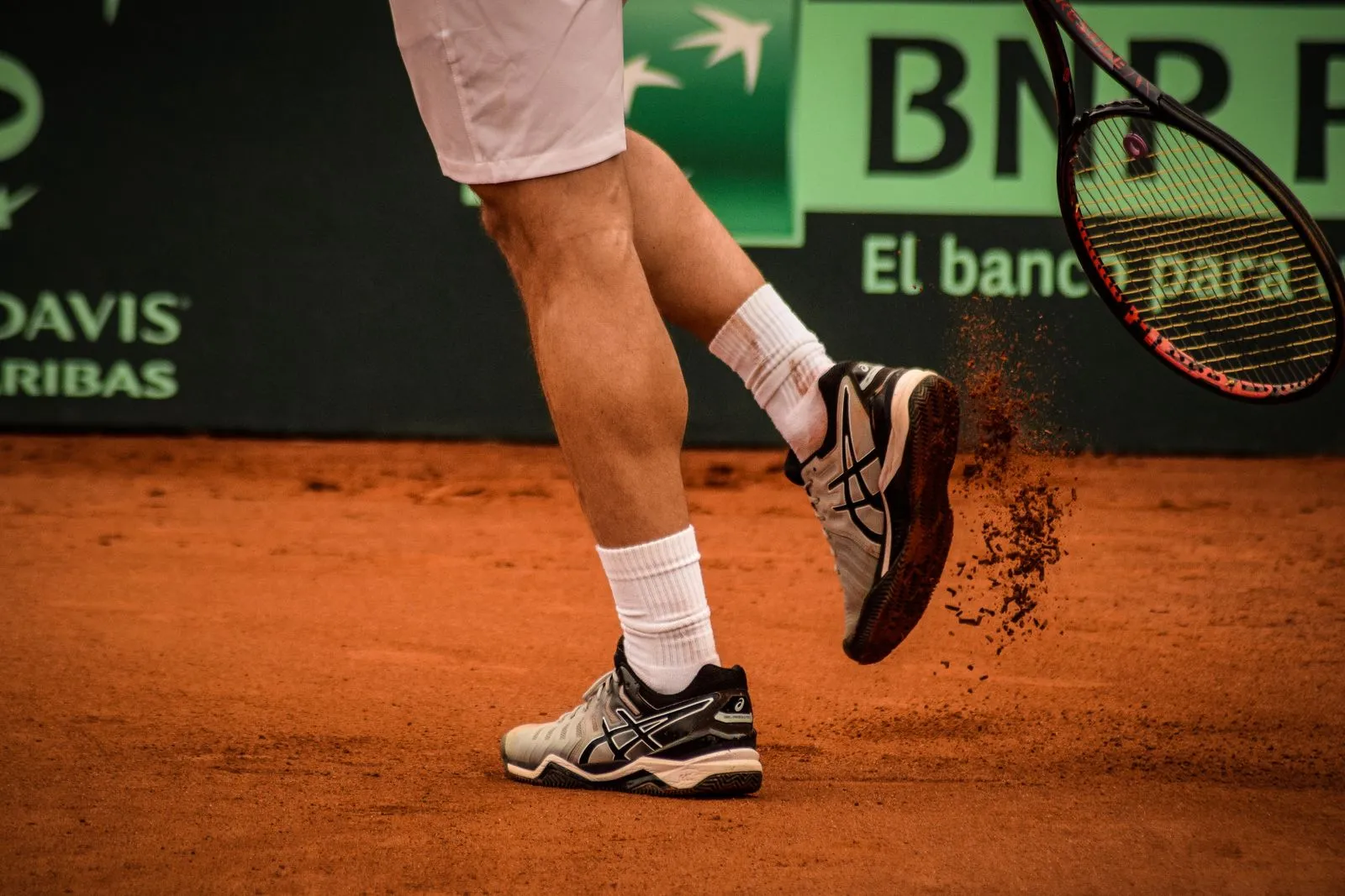 Jugador de tenis en movimiento sobre polvo de ladrillo, con un banner de sponsor de fondo